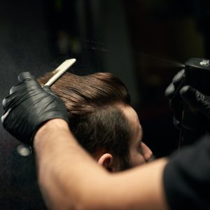 Crop of hairdresser's hands in black gloves combing clean male client hair with hairbrush and sprinkle it with water.Handsome man getting new hairstyle at modern and professional salon